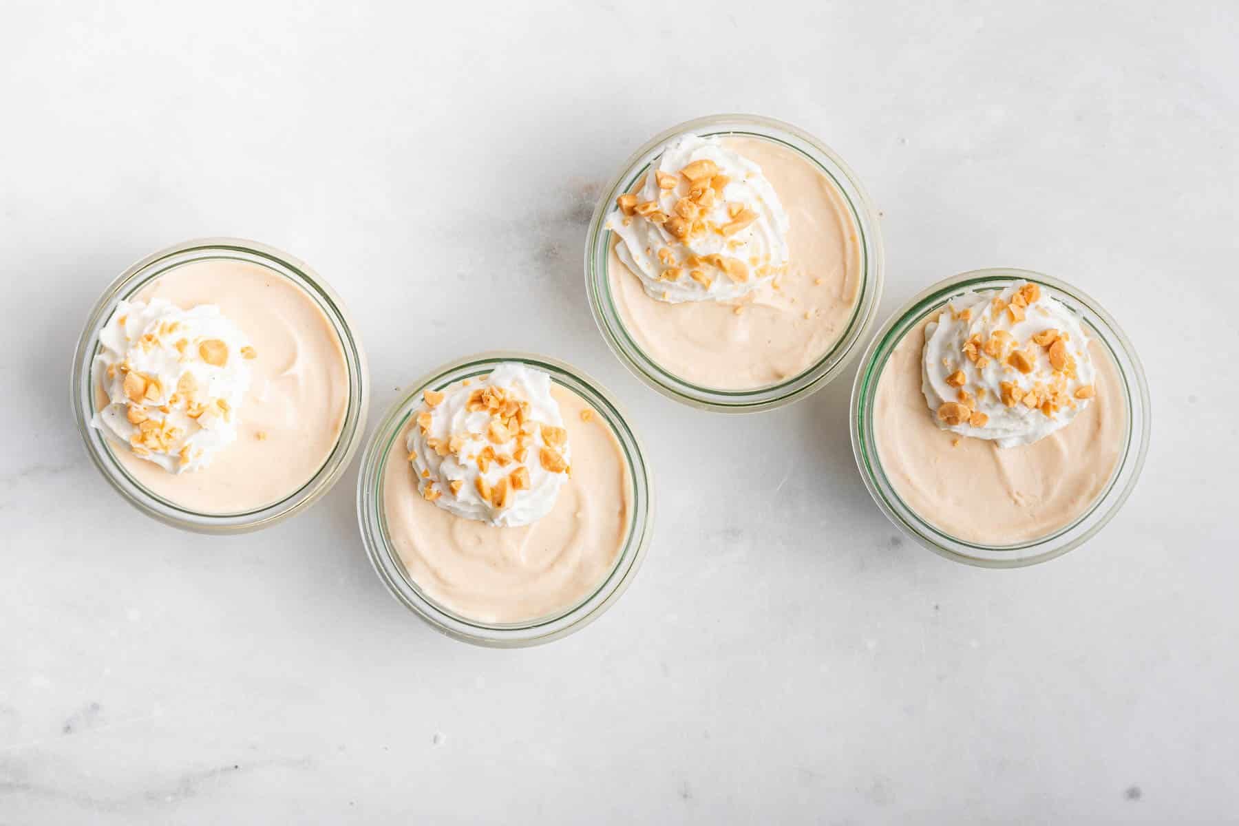 Horizontal image of 4 bowls of a light brown dessert with whipped cream and peanuts on top.