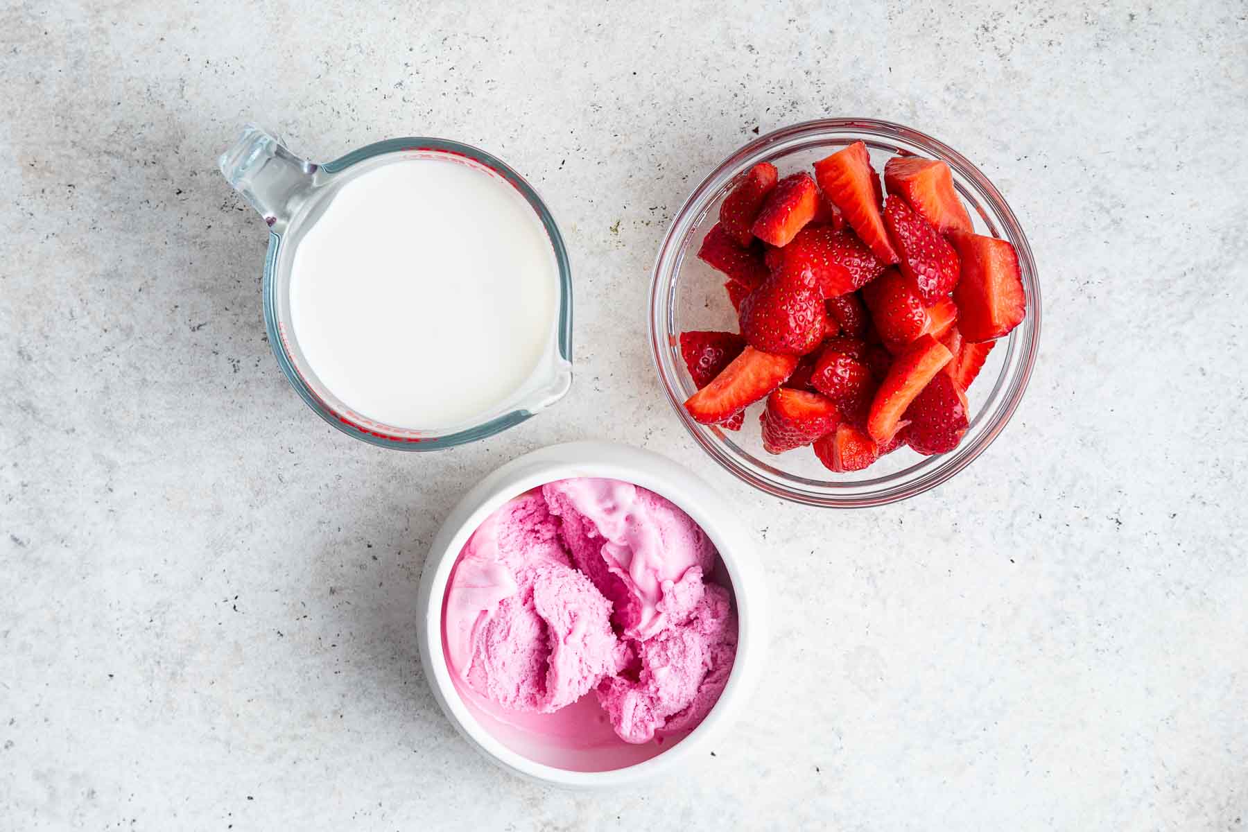 Schüssel mit frischen Erdbeeren, Schüssel mit rosa Erdbeereis und Messbecher mit Milch auf der Theke.