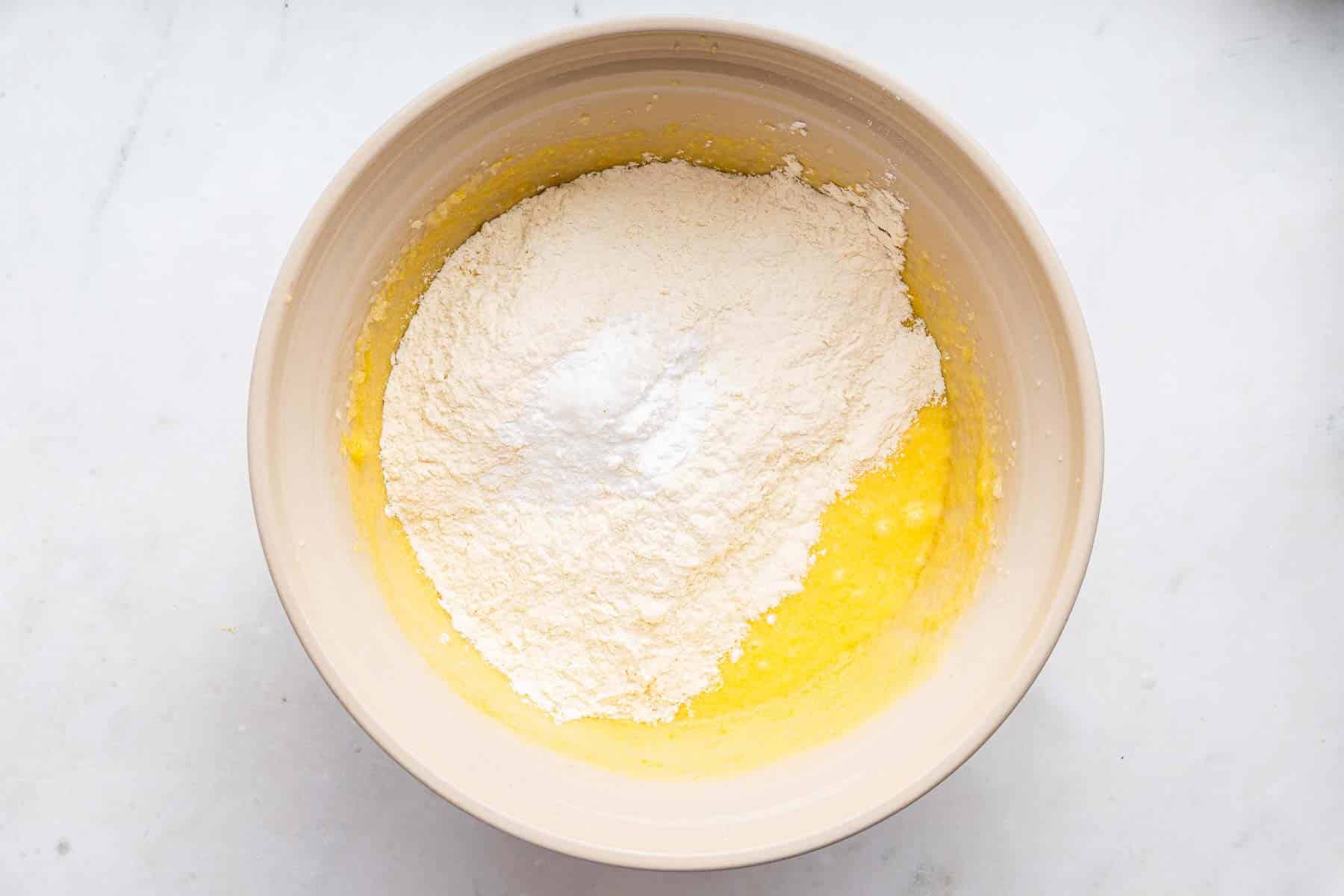White flour on top of yellow batter in cream bowl on counter.